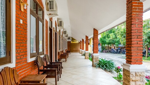 A covered outdoor seating area with brick walls and tiled flooring, featuring rows of wooden tables and chairs. - RedDoorz Syariah near Gor Ken Arok (Malang)