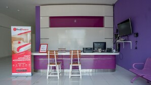 A reception area with a purple and white color scheme, a desk with a computer, two wooden chairs, and a large RedDoorz advertisement. - RedDoorz near Alun Alun Kaliwungu Kendal (Kaliwungu)