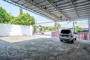 Un área de estacionamiento techada con una camioneta blanca estacionada bajo un techo metálico, rodeada de pavimento y una valla.