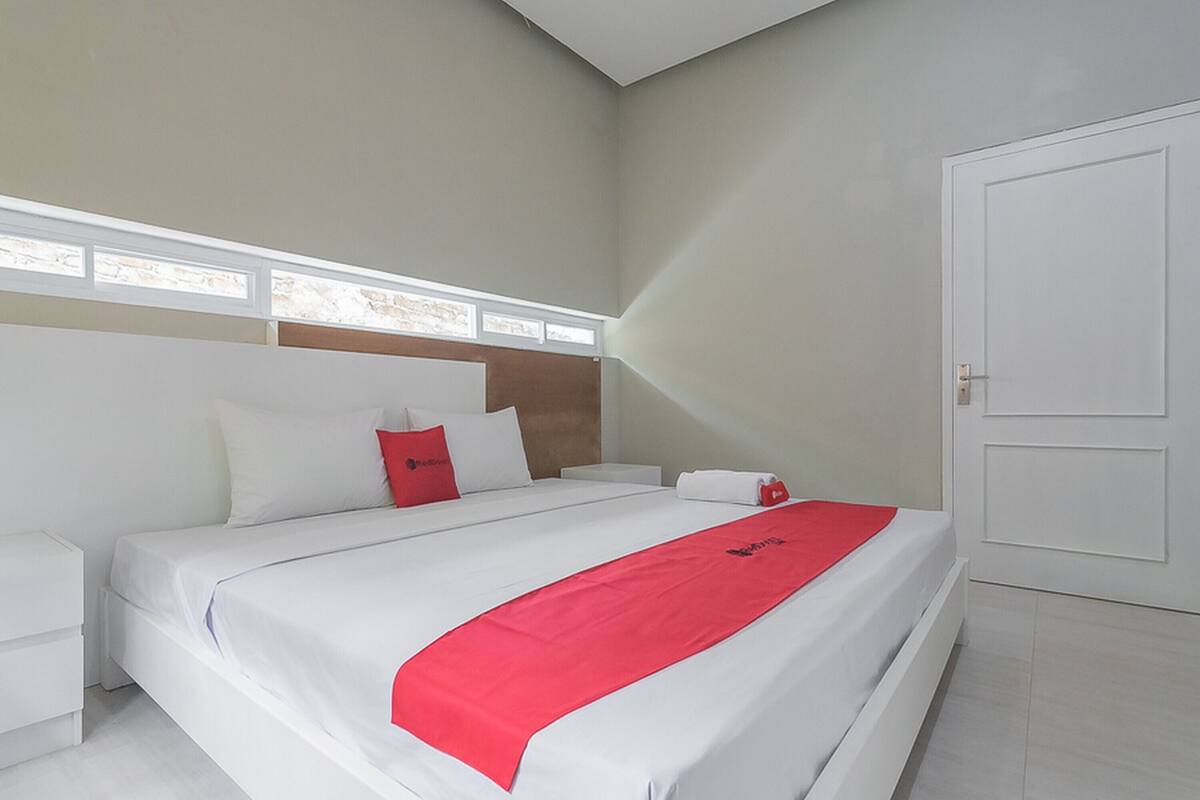 A modern hotel room with a large bed, white bedding, a red accent pillow, and a white nightstand.
