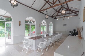 A spacious dining area with a long wooden table, white chairs, and large windows allowing natural light. - RedDoorz Resort Syariah @ Idelansia Homestay Ciater (Ciater)