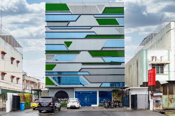 Un edificio moderno con fachada geométrica, entrada azul y estacionamiento con coches y motocicletas.
