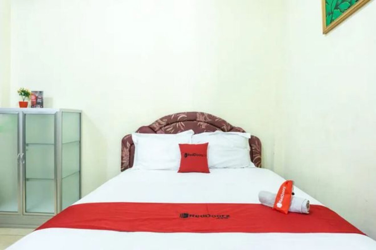 A neatly made bed with a red blanket, a pillow with a logo, and a rolled towel on the side.