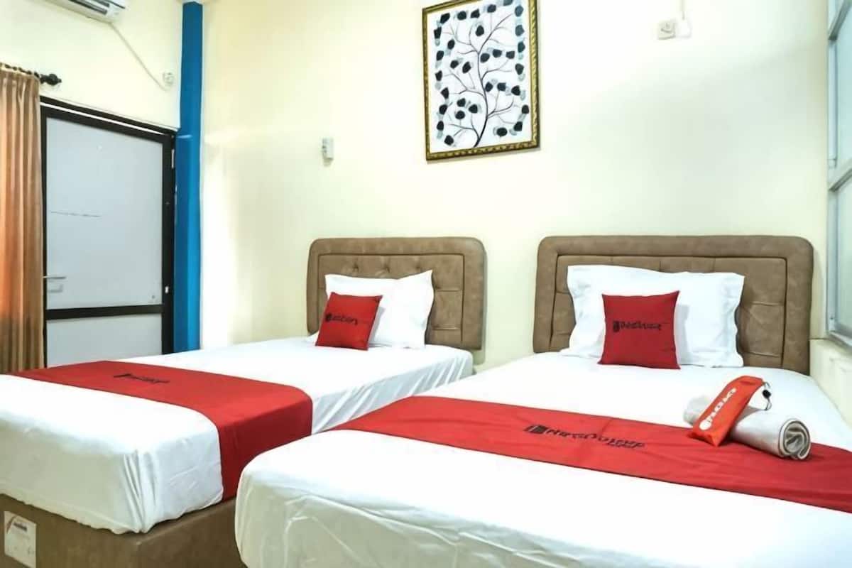 Two beds with red bedspreads, a blue pillar, a door, and a framed artwork on the wall.