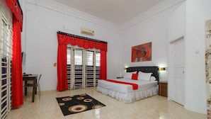 Un dormitorio con cama, escritorio, silla, ventana con cortinas rojas y un cuadro en la pared.