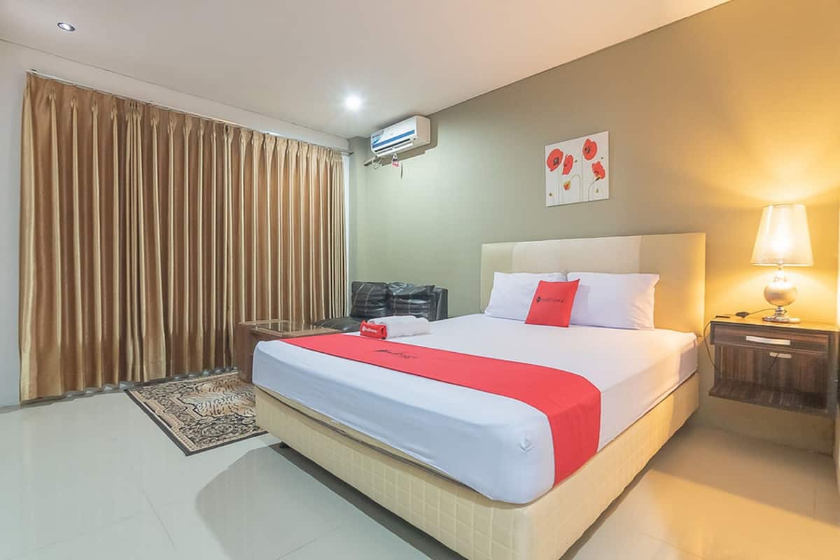 Habitación de hotel con cama, mesita de noche, lámpara, unidad de aire acondicionado, ventana con cortinas y una obra de arte en la pared.