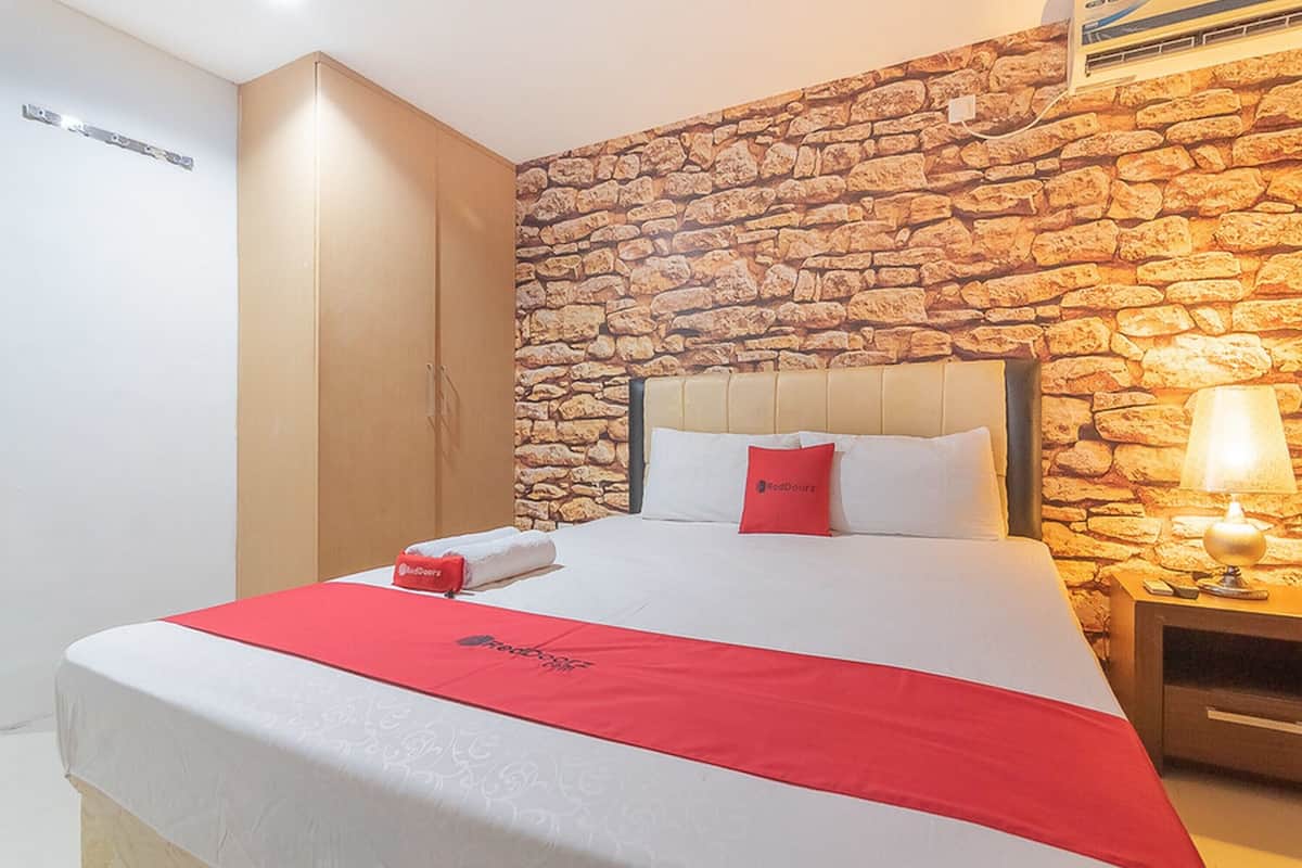 Habitación de hotel con pared de piedra, cama con ropa de cama blanca y detalles rojos, armario y mesita de noche con lámpara.