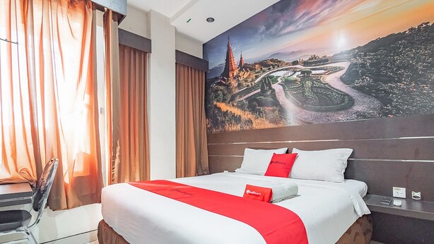A hotel room with a large bed, a desk, and a chair. There is a painting on the wall depicting a landscape. - RedDoorz near Stadion Sanaman Mantikei (Palangkaraya)
