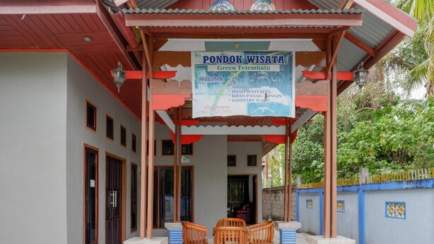 Área externa coberta com uma placa indicando 'Pondok Wisata Green Tetembatu', listando comodidades como chuveiro, área de lavagem das mãos e sanitários. Há três cadeiras de madeira e uma mesa sob a proteção.