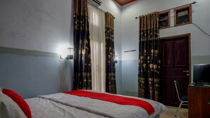 Quarto com cama, escrivaninha e cadeira. Há uma janela com cortinas, uma porta e ar-condicionado fixado na parede.