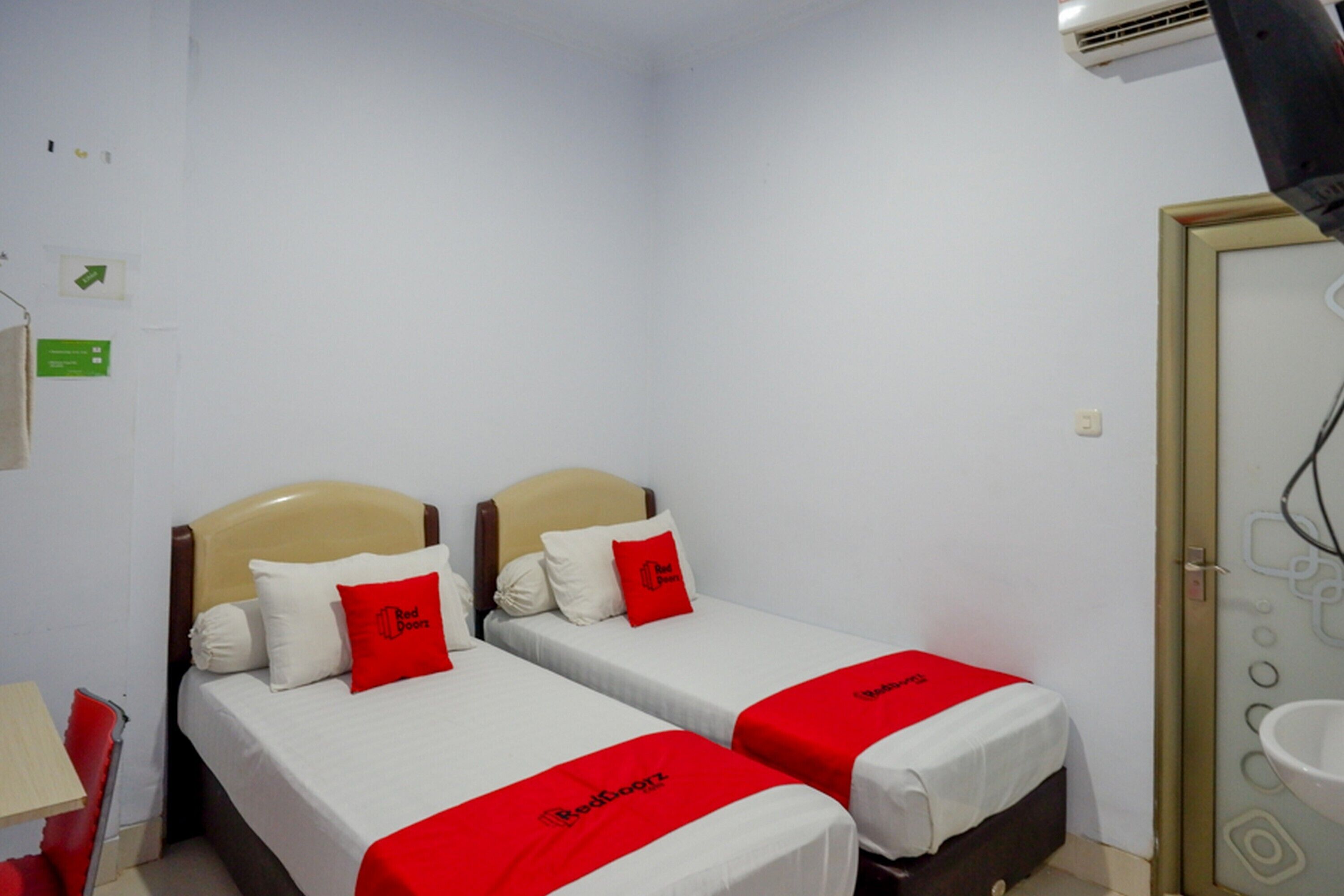 Habitación de hotel con dos camas, cada una con colcha roja y almohada con el logo del hotel. Se ve una pared blanca, una puerta con panel de vidrio y una mesita de noche con una lámpara.