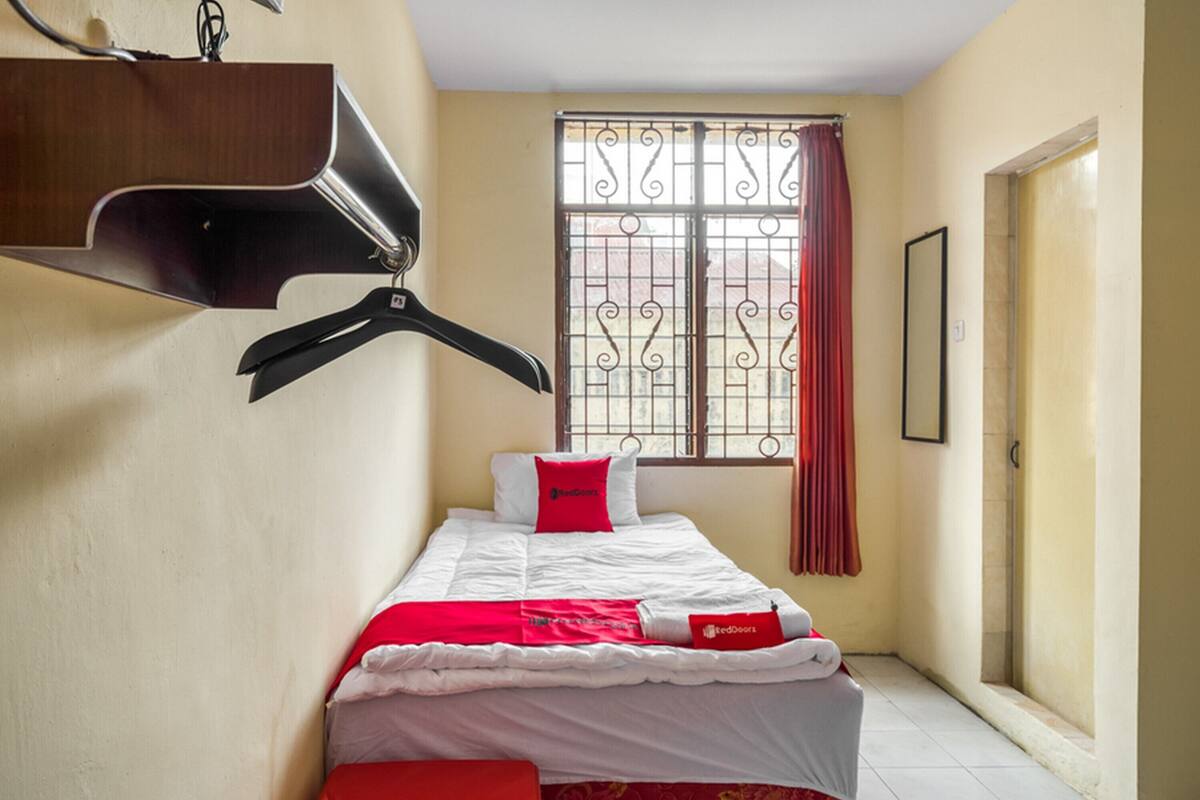 A small, neatly arranged bedroom with a bed, a hanging coat rack, a window with bars, and a door.