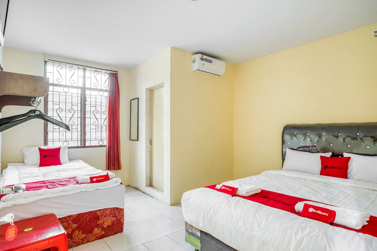A hotel room with two beds, a red and white color scheme, an air conditioning unit, and a window with decorative bars.