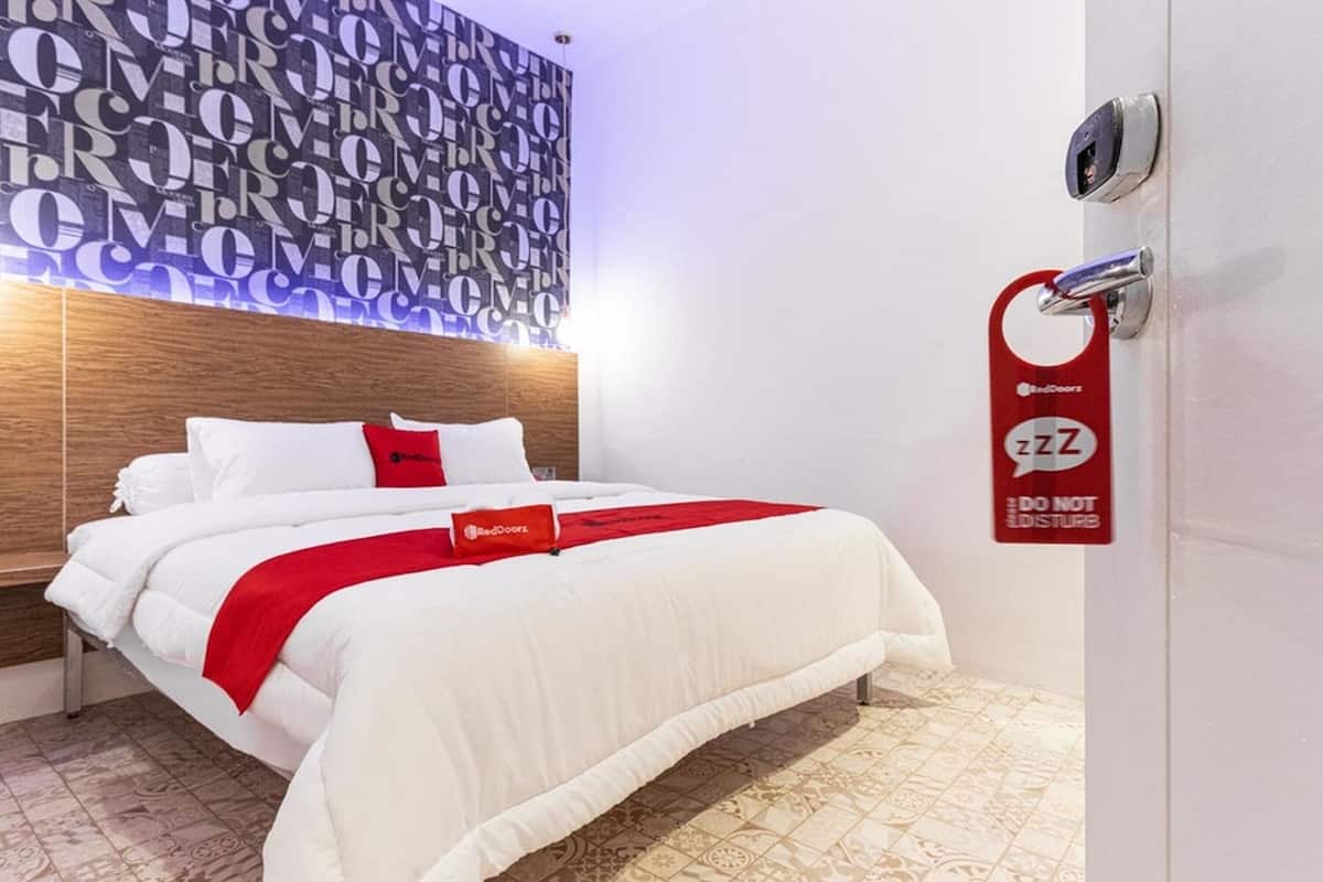 A hotel room with a bed, a red and white bedspread, a patterned wall, and a door with a red sign.
