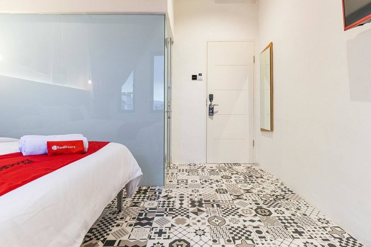 A hotel room with a bed, a red towel, a RedDoorz sign, a patterned floor, a door, and a mirror on the wall.