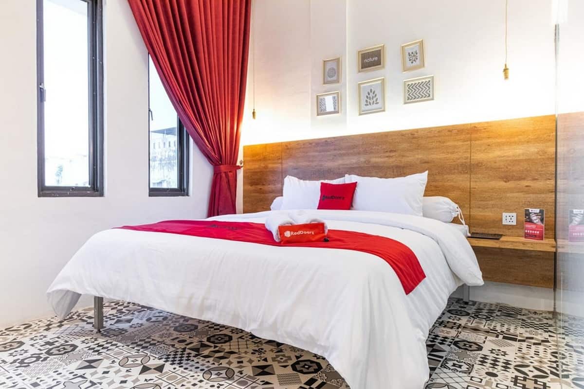 A hotel room with a bed, a red curtain, a wooden headboard, and patterned floor tiles.
