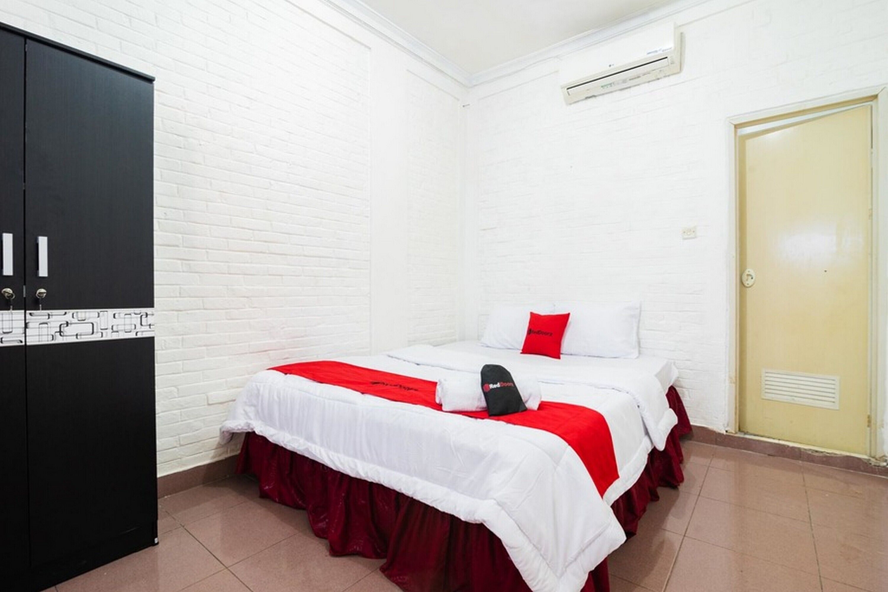 A hotel room with a white brick wall, a bed with red bedding, a black wardrobe, and a door with a vent.