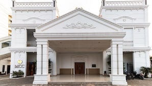 A white building with a central entrance flanked by two wings, a sign reading "MCC" on the left, and a paved area in front.