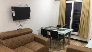 Una sala de estar con un sofá marrón, una mesa de comedor de vidrio con sillas, un televisor de pantalla plana montado en la pared y una ventana con cortinas.