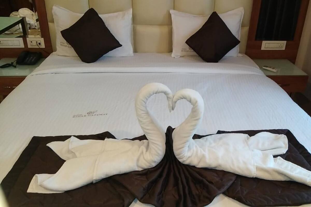 Habitación de hotel con una cama bien hecha, una toalla dispuesta en forma de corazón y un minibar.