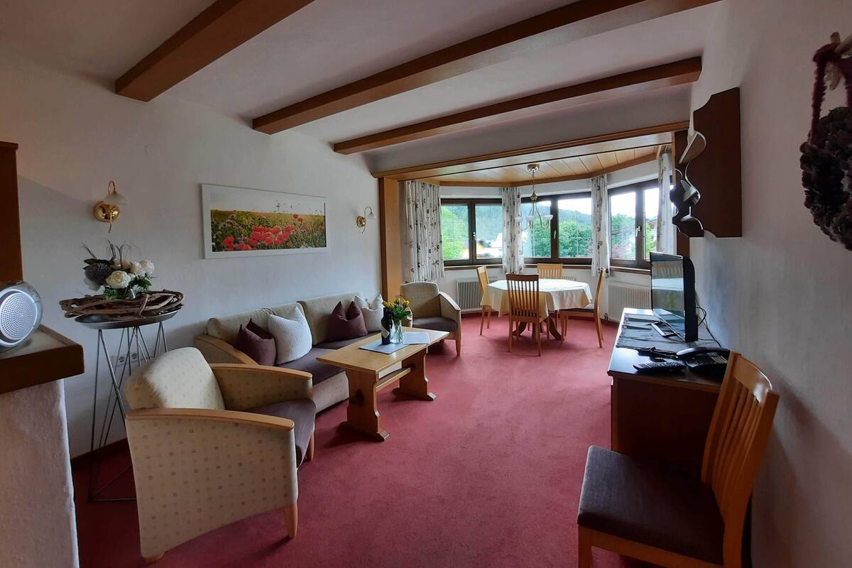 A cozy living room with a sofa, chairs, a coffee table, and a dining table. There are windows with curtains, a wall-mounted lamp, and a speaker.