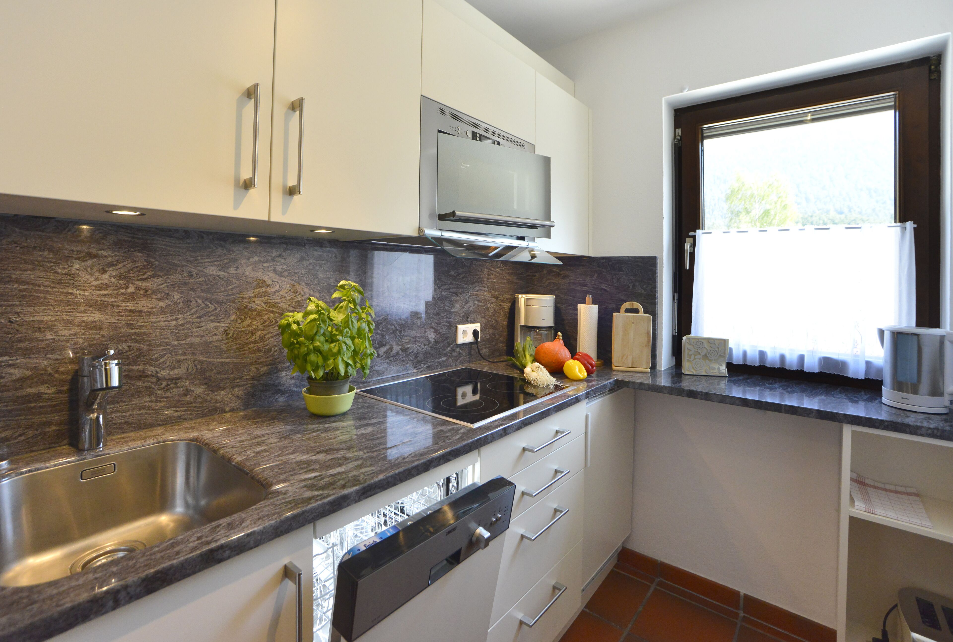 A modern kitchen with a stainless steel sink, a built-in oven, and a window with a view of greenery.