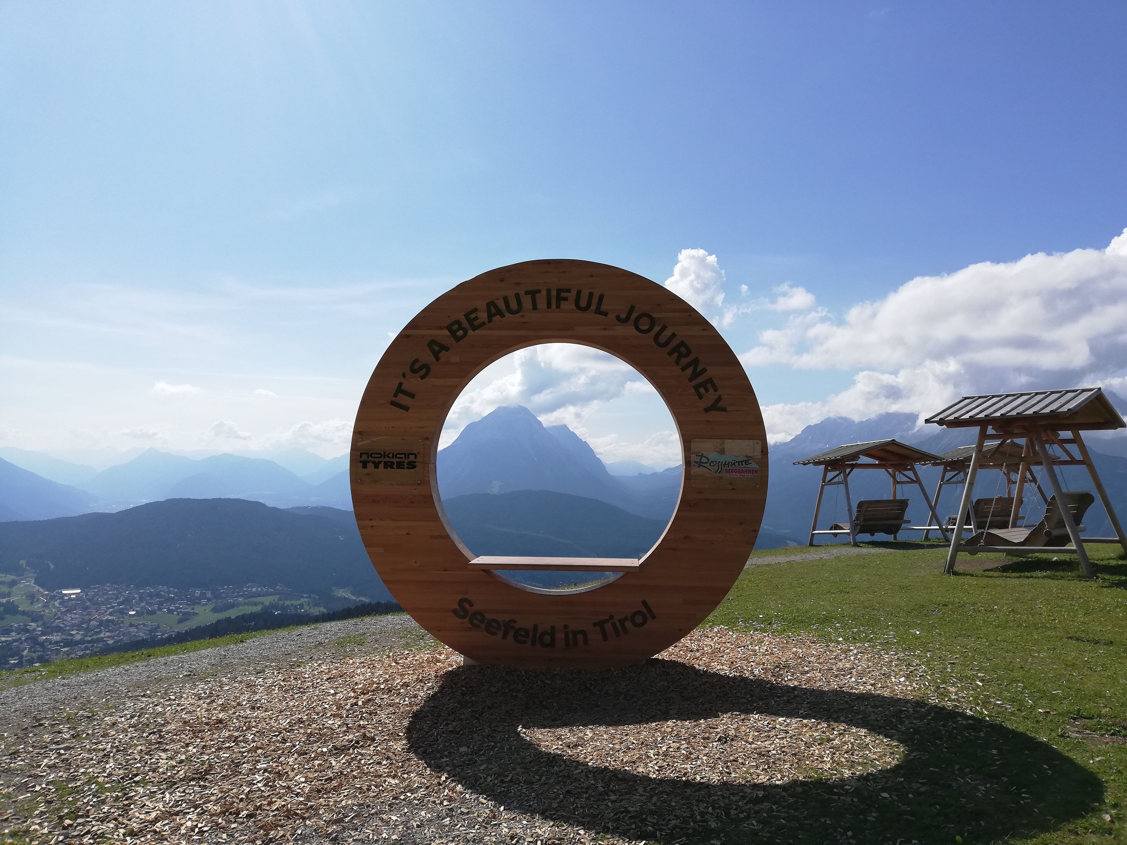 A wooden sign with "It's a beautiful journey" and "Seefeld in Tirol" text, set against a mountainous backdrop with a clear sky.