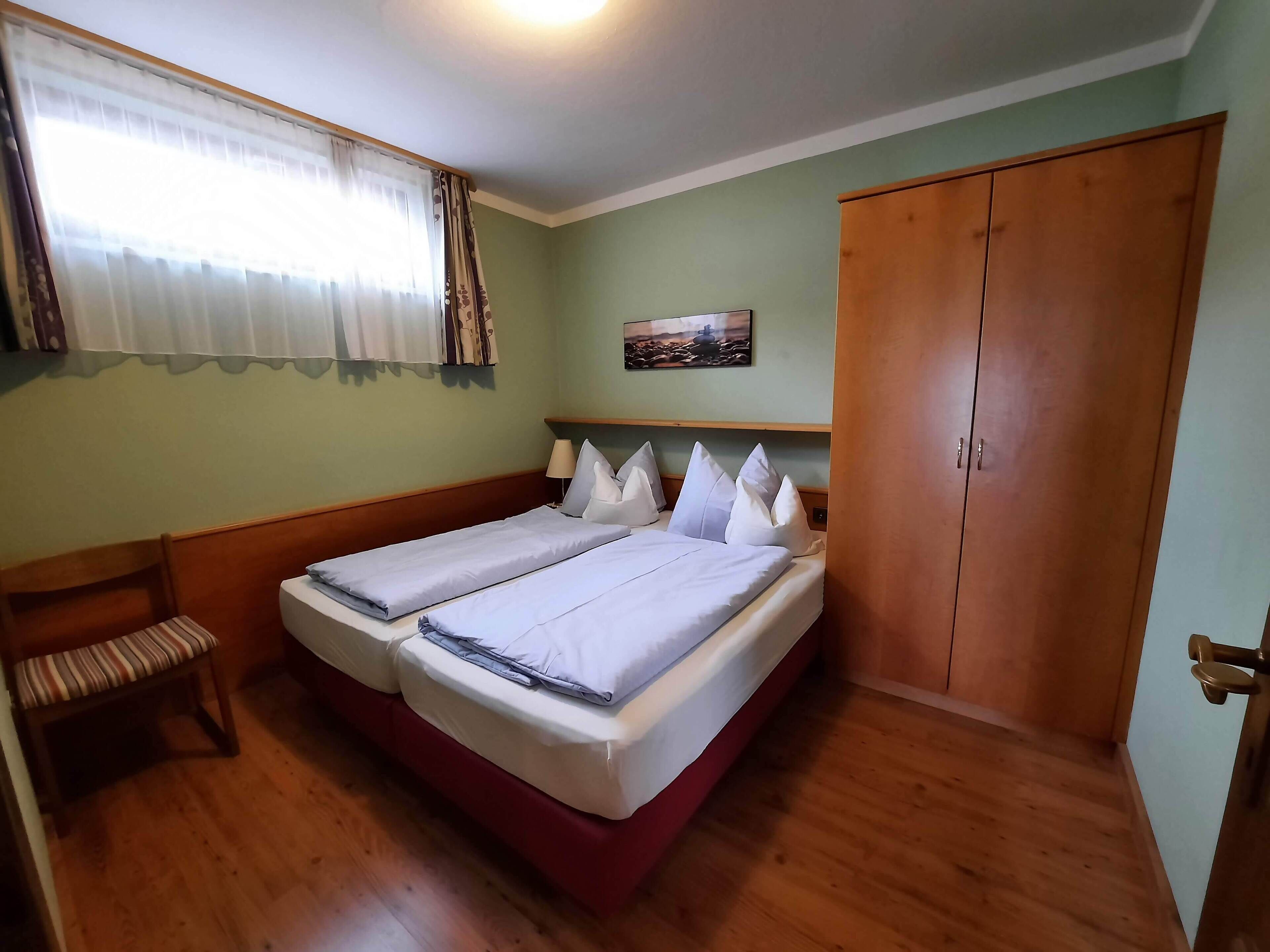 Une chambre avec deux lits, un banc en bois, une armoire et une fenêtre avec des rideaux.