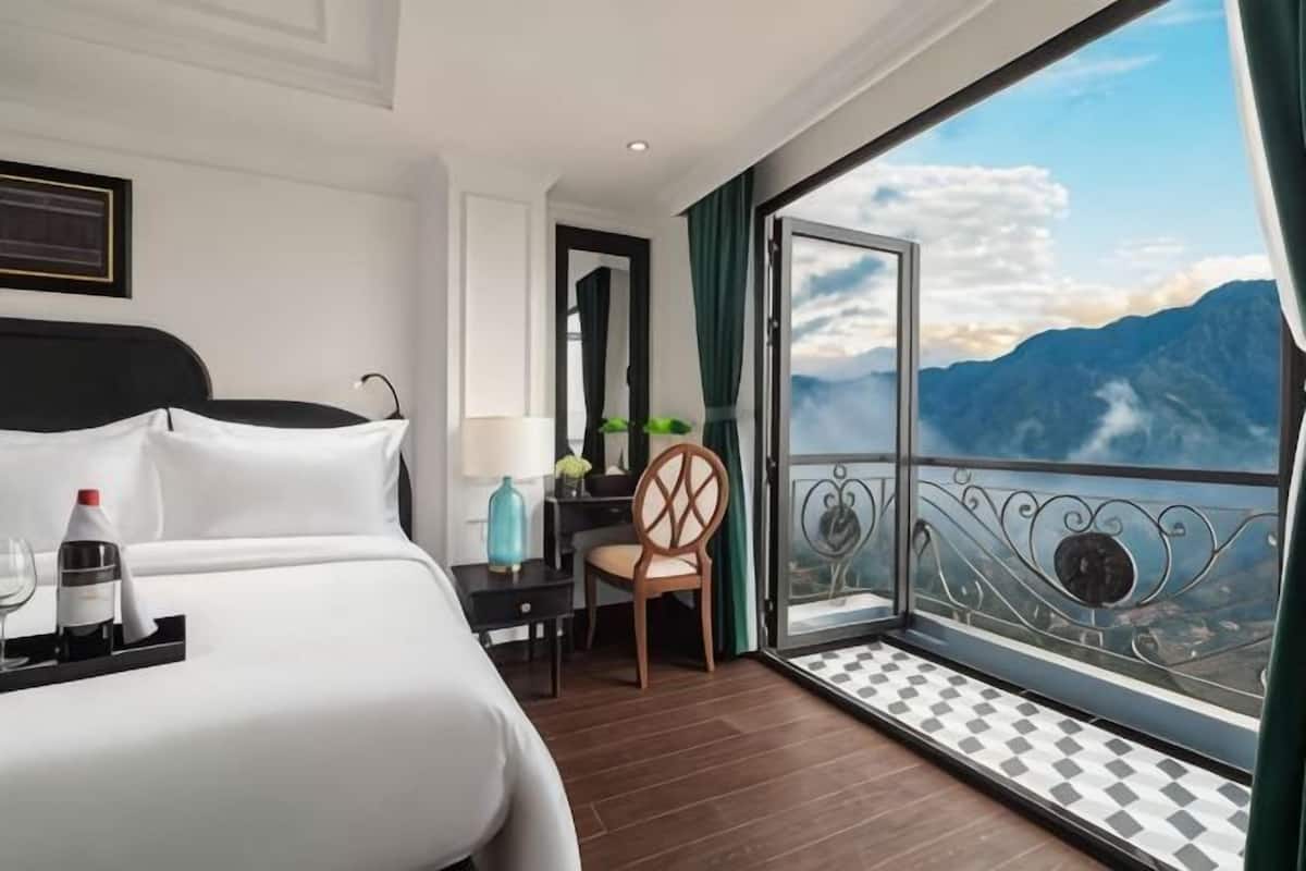 A hotel room with a bed, a chair, a table with a lamp, a tray with a bottle and glasses, and a balcony with a view of mountains.