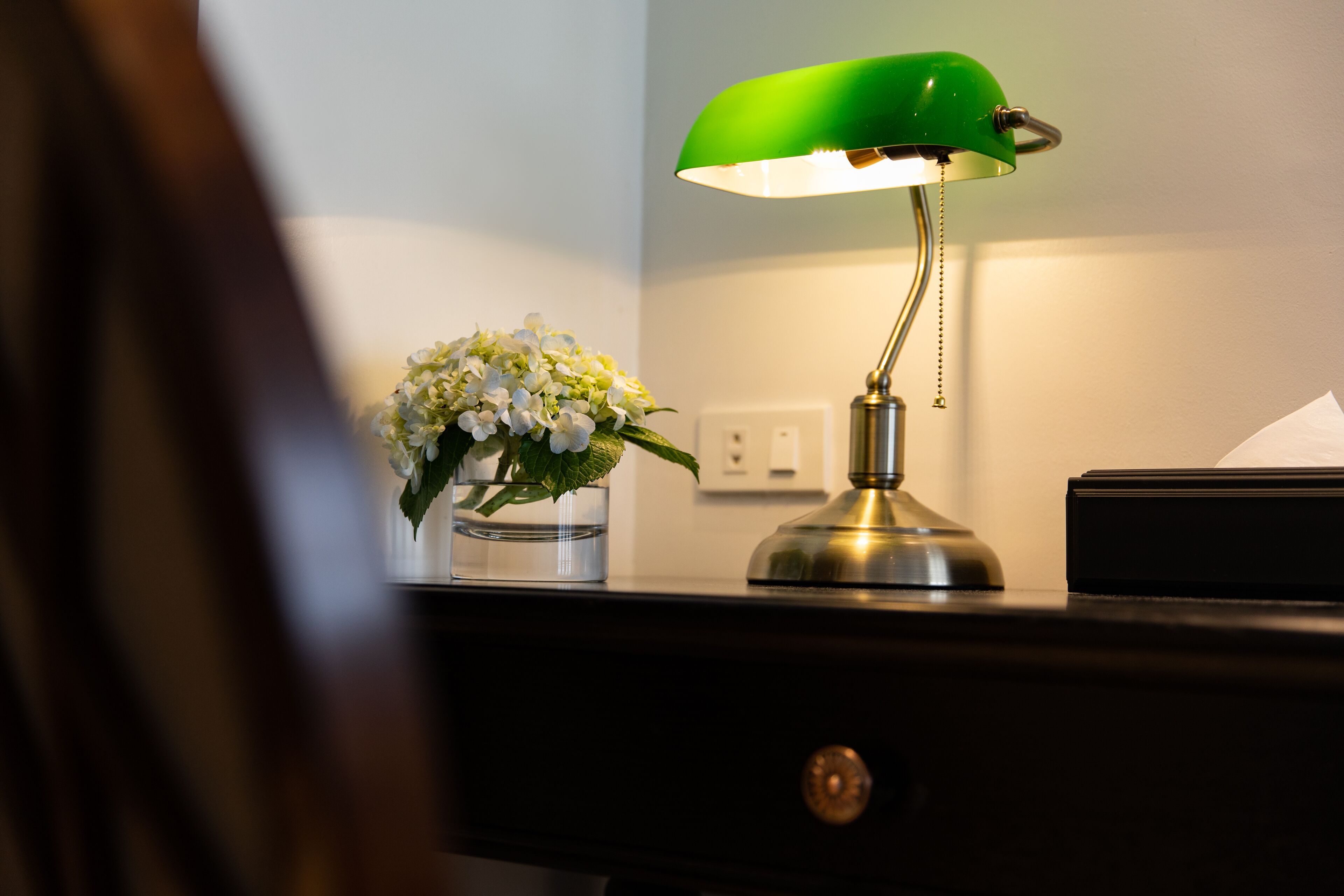 Un escritorio con una lámpara de escritorio verde, un jarrón con flores blancas y una caja negra.