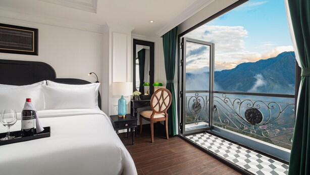 A hotel room with a large bed, a chair, a small table with a bottle and glasses, and a balcony with a mountain view. - My Gallery Boutique Hotel & Spa (Sa Pa)