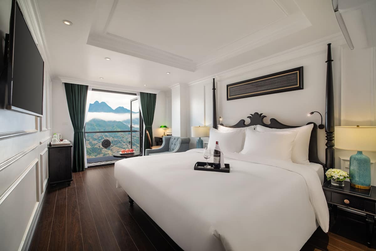 A hotel room with a large bed, a nightstand, a TV, and a window with a view of mountains.