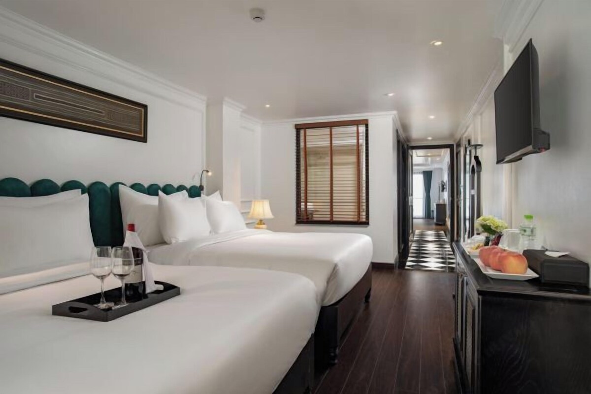 Quarto de hotel com duas camas, televisão fixada na parede, bandeja com copos de vinho e piso xadrez.