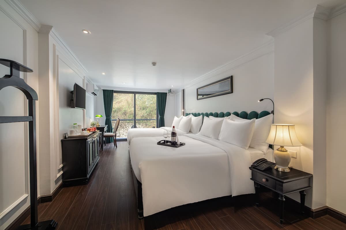 A hotel room with two beds, a desk with a chair, a TV, and a window with a view of greenery.