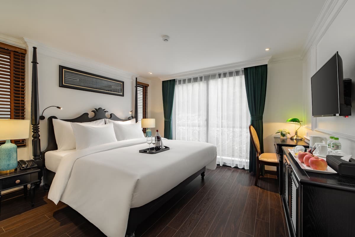 A hotel room with a large bed, a desk with a chair, a flat-screen TV, and a window with curtains.