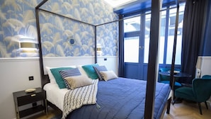 A four-poster bed with a patterned bedspread, a bedside table with a lamp, and a view of the outdoors through a window. - Le Cafe des Invalides - Eiffel Tower (Paris)