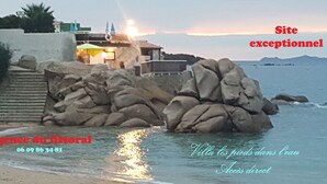En fastighet vid kusten med en villa som ligger delvis i vattnet, en strand och en trappa som leder upp till en byggnad.