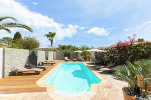 A rectangular swimming pool with a stone border, surrounded by lounge chairs and umbrellas, set against a stone wall and lush greenery.