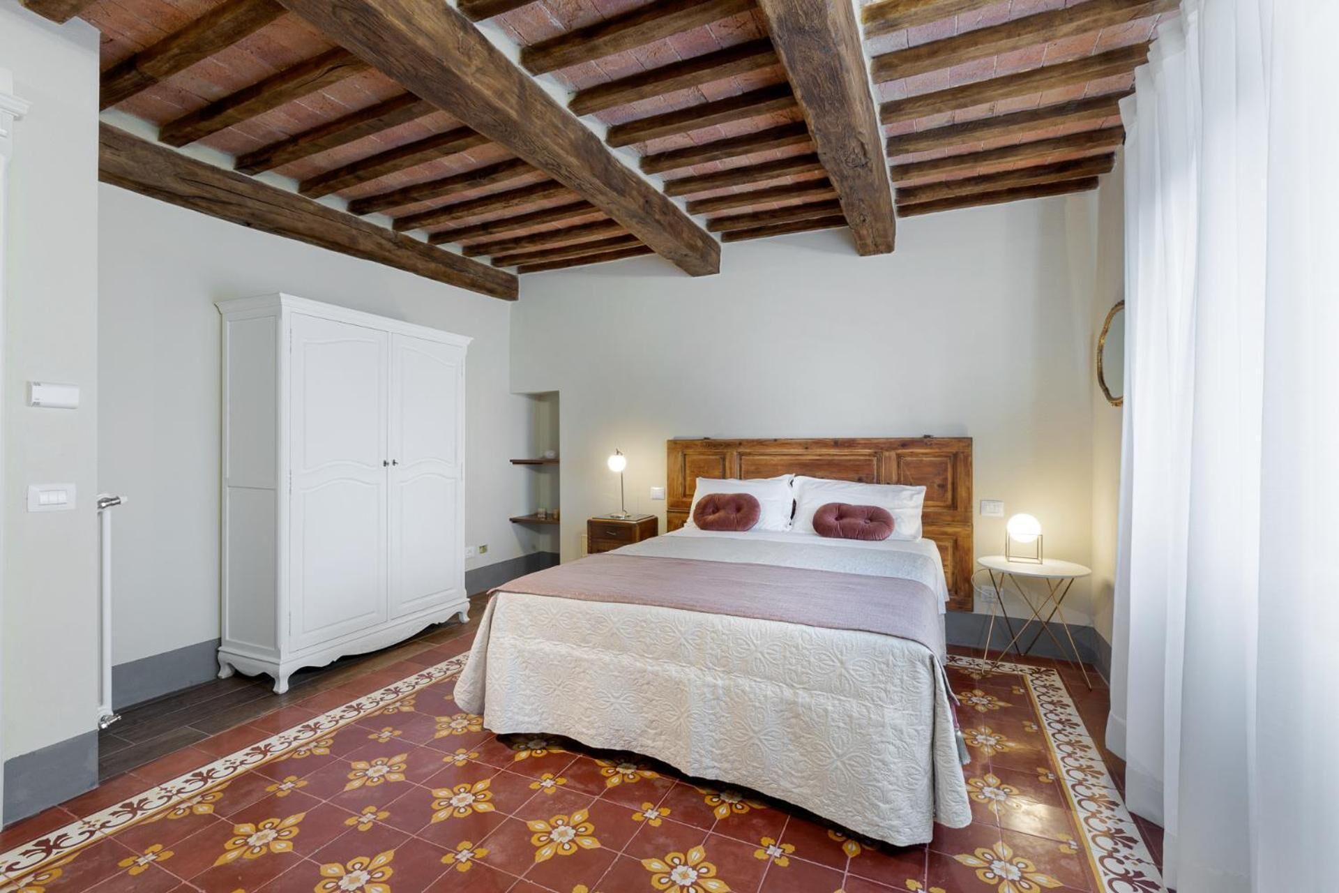 Un dormitorio con techo de madera, una cama con colcha blanca y marrón, un armario blanco y una mesita de noche con lámpara.