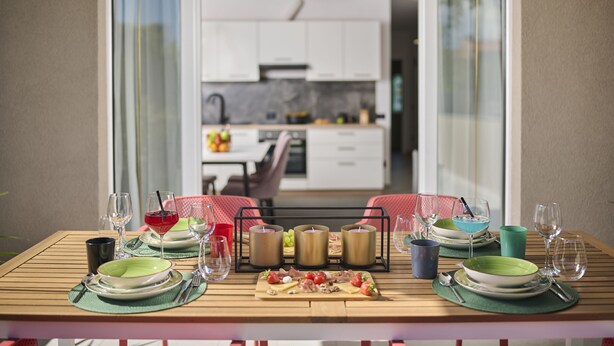 Mesa de jantar posta com pratos, copos e velas, com vista para uma cozinha ao fundo.