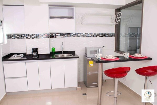 A compact kitchen with white cabinets, a black countertop, a microwave, a coffee maker, and a sink. There are two red bar stools and a small window above the sink.