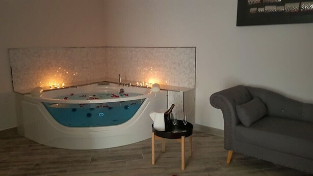 Habitación de esquina con bañera, una mesita con una botella y un vaso, y un sofá gris.