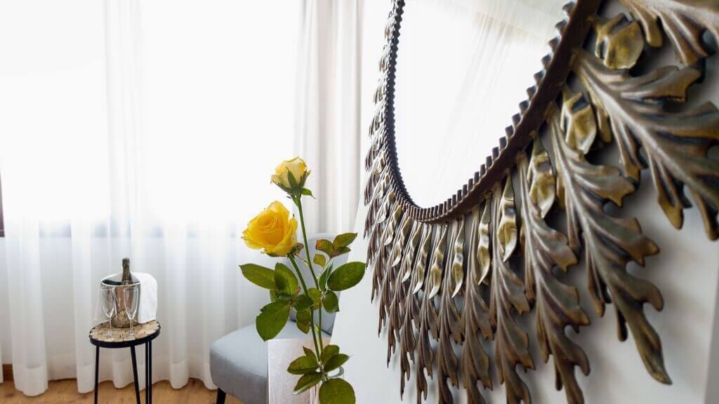 A room with a decorative mirror, a vase with yellow flowers, and a small table with a glass object.