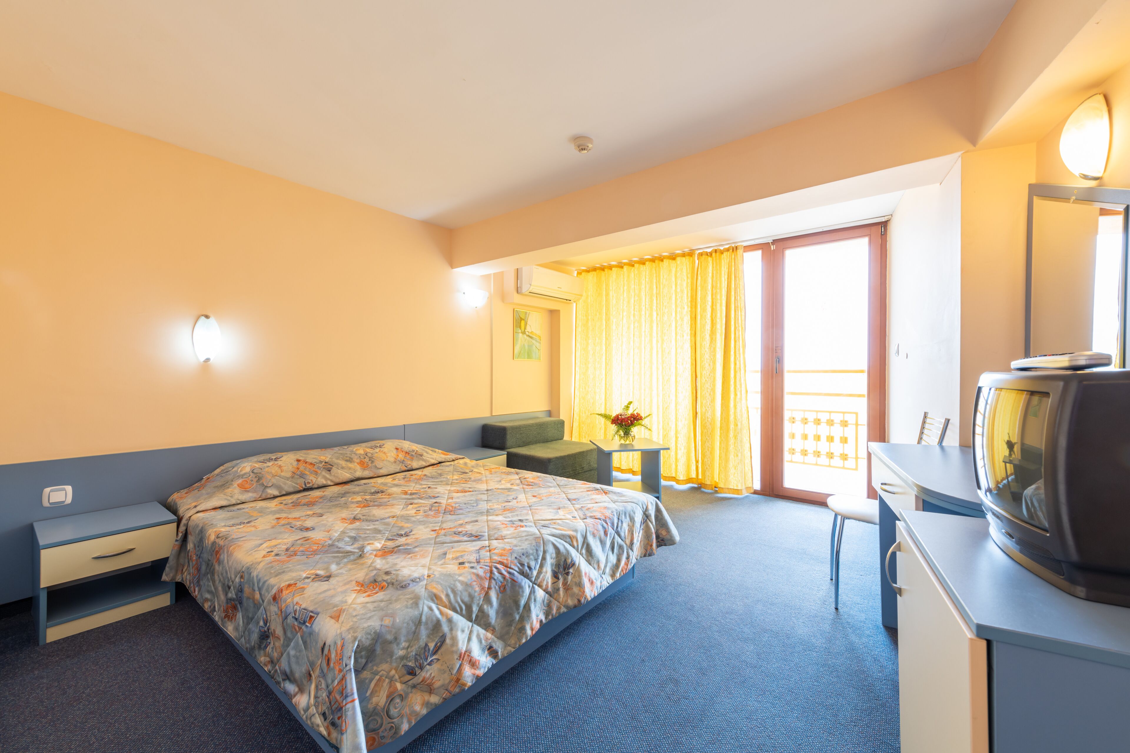 Habitación de hotel con cama, escritorio con televisor, silla, mesita con florero y balcón con cortinas amarillas.