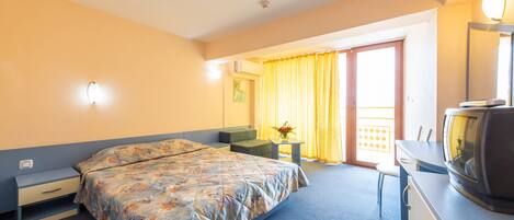 Habitación de hotel con cama, escritorio con televisor, silla, mesita con florero y balcón con cortinas amarillas.