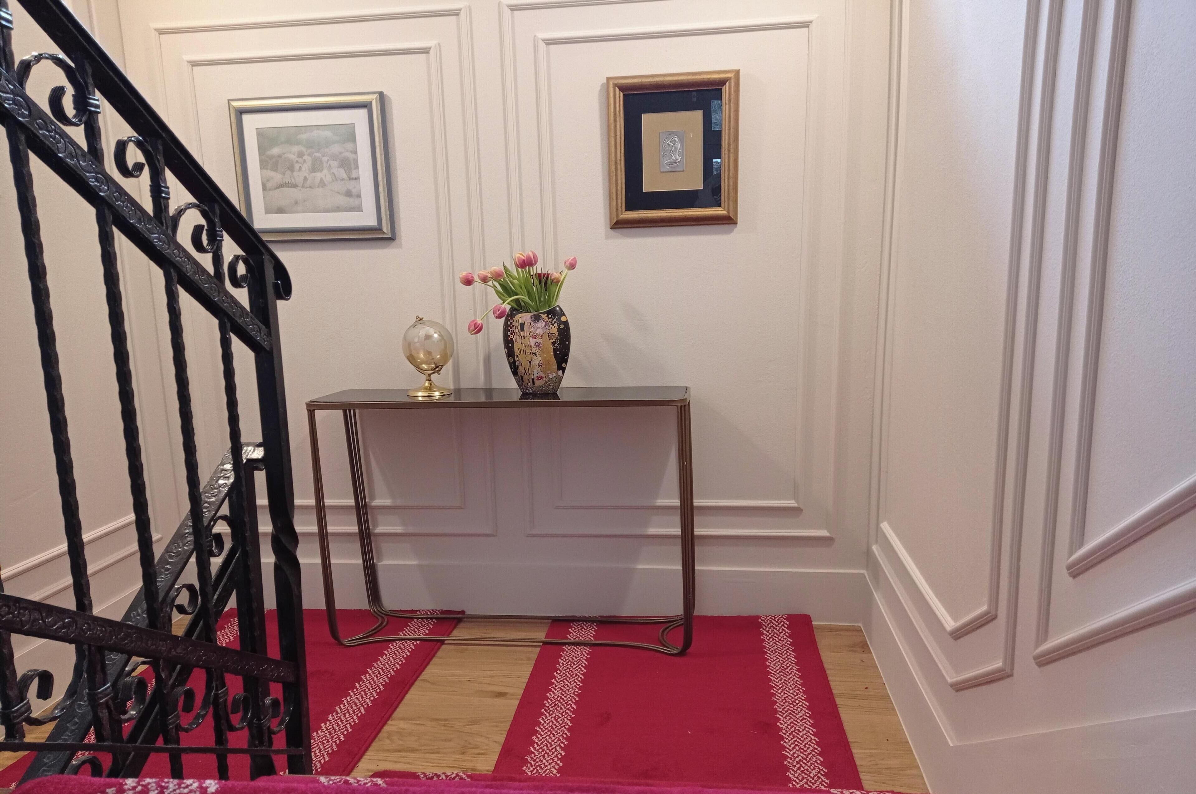 Een trap met een rood tapijt, een console tafel met een bloevas en lijstjes aan de muur.