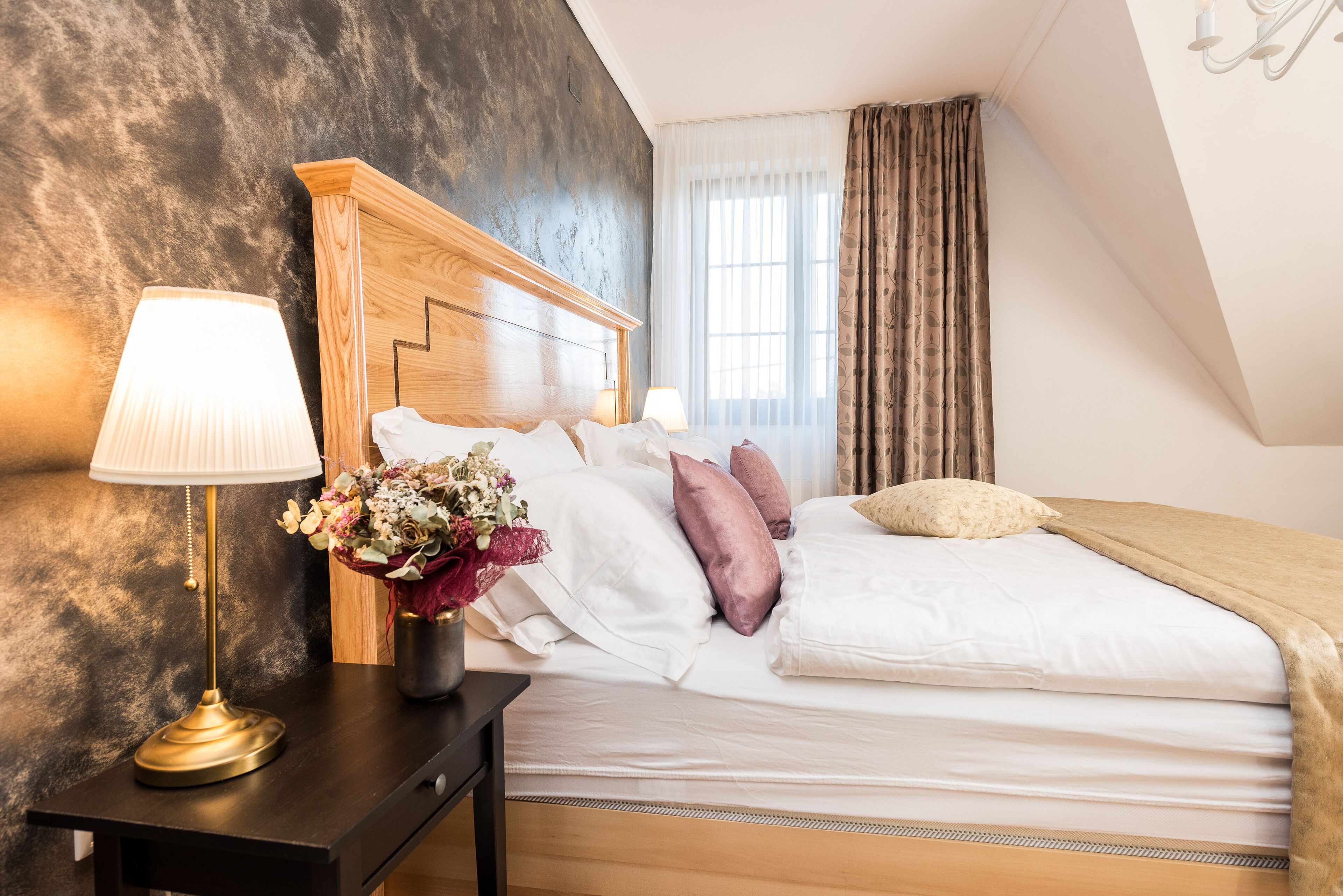 A neatly made bed with white and pink pillows, a bedside lamp, and a vase of flowers on a wooden nightstand.