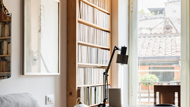 Una habitación acogedora con una estantería, un escritorio con lámpara y una ventana con vistas a edificios.