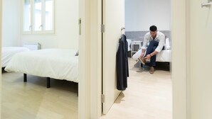Hotellihuone, jossa on sänky, pieni pöytä ja henkilö istuu lattialla ja sitoo kengännauhojaan.
