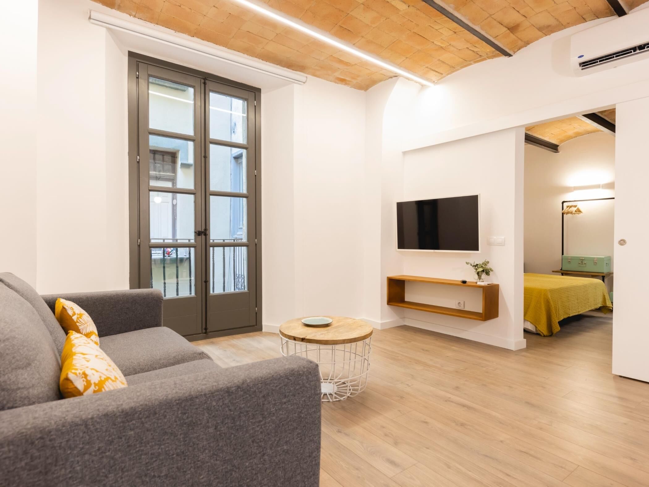 A modern living room with a grey sofa, a small round coffee table, a TV mounted on the wall, and a wooden ceiling.